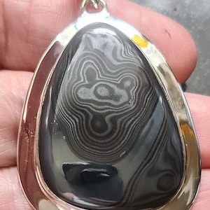 Puede incluir: Colgante en forma de lágrima con una piedra pulida de color gris oscuro que presenta un patrón único de líneas grises claras. La piedra está engastada en un marco plateado pulido con una argolla a juego.