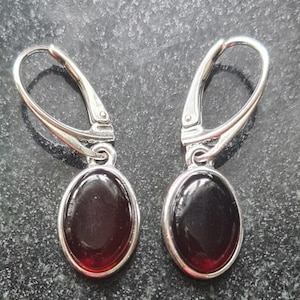 May include: A pair of silver leverback earrings with oval, dark red gemstones. The gemstones are set in a silver bezel. The earrings are displayed on a dark, textured surface.