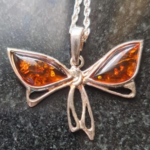 May include: A silver-toned pendant necklace featuring a butterfly design. The butterfly wings are filled with amber-colored resin, and the necklace has a delicate chain. The pendant is detailed and has a polished finish.