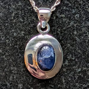 May include: A silver pendant necklace with an oval, dark blue gemstone set in a polished silver frame. The pendant is suspended from a delicate, twisted silver chain. The background is a dark, textured surface.