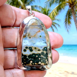 May include: A polished silver pendant featuring a large, shield-shaped ocean jasper gemstone. The stone displays a gradient of colours, transitioning from a dark forest green base with circular orb patterns to lighter blue, white, and tan bands.