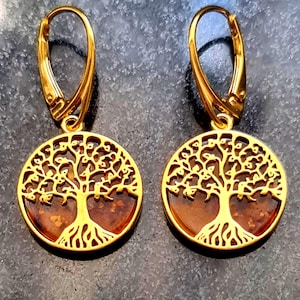 May include: A pair of gold-colored earrings featuring a tree of life design. The earrings have a circular pendant with a brown center and a detailed tree silhouette. The lever-back closures are also gold-colored.