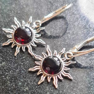 May include: A pair of silver-toned earrings featuring a sunburst design. Each earring has a central, round, dark red gemstone. The sunburst has pointed rays extending outwards. The earrings have a lever-back closure.