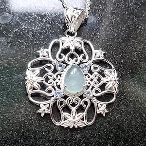 Puede incluir: Un collar con colgante de plata con un diseño floral. El colgante presenta una piedra preciosa azul pálido en forma de pera en el centro, rodeada de piedras preciosas azules más pequeñas y filigrana plateada. El collar está en una cadena de plata.
