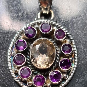 Puede incluir: Colgante de plata con una gema ovalada de color marrón claro en el centro, rodeada de diez gemas redondas moradas más pequeñas. El colgante tiene un borde en forma de cuerda y una argolla de plata.