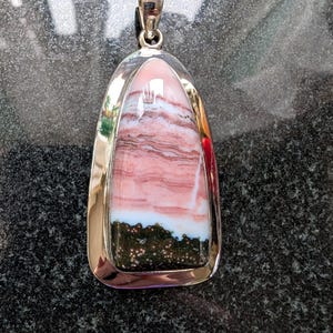 May include: A teardrop-shaped pendant featuring a polished stone with horizontal bands of pink and white, transitioning into a dark green base with small white speckles. The stone is encased in a smooth, polished silver-toned metal setting.