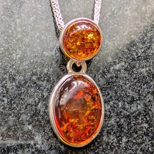 May include: A silver pendant necklace featuring two amber-colored, oval and round, cabochon stones. The stones are set in silver bezels and connected by a silver loop. The necklace chain is a silver curb chain.