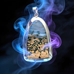 May include: A silver pendant featuring an oval-shaped ocean jasper stone with distinct circular patterns in shades of tan, green, and white. The stone is encased in a polished silver frame with a bail at the top, surrounded by swirling blue and purple smoke.