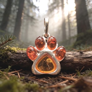 May include: A silver pendant in the shape of a paw print, featuring amber-colored stones. The pendant is set against a blurred forest background, with sunlight filtering through the trees. The paw print design is a popular theme for pet lovers.