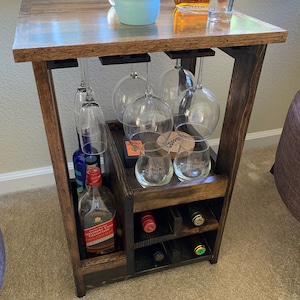 May include: A dark stained wooden bar cart with a tabletop, wine glass holders, and wine bottle storage. The cart holds wine glasses, bottles of liquor, and a small shot glass. The cart is designed for home entertaining.
