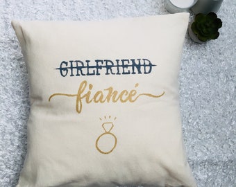 Puede incluir: Cojín color crema con las palabras "GIRLFRIEND" tachadas y reemplazadas por "fiancé" en letras doradas. Un dibujo de un anillo dorado está debajo. El cojín está sobre una superficie blanca texturizada.