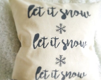 Może przedstawiać: Biała poduszka ozdobna z napisem "Let it snow" powtórzonym trzykrotnie czarnym pismem. Każdy wiersz tekstu jest ozdobiony czarną śnieżynką.