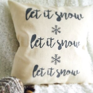 May include: A white throw pillow with the text "Let it snow" repeated three times in black lettering. Each line of text is accompanied by a black snowflake.