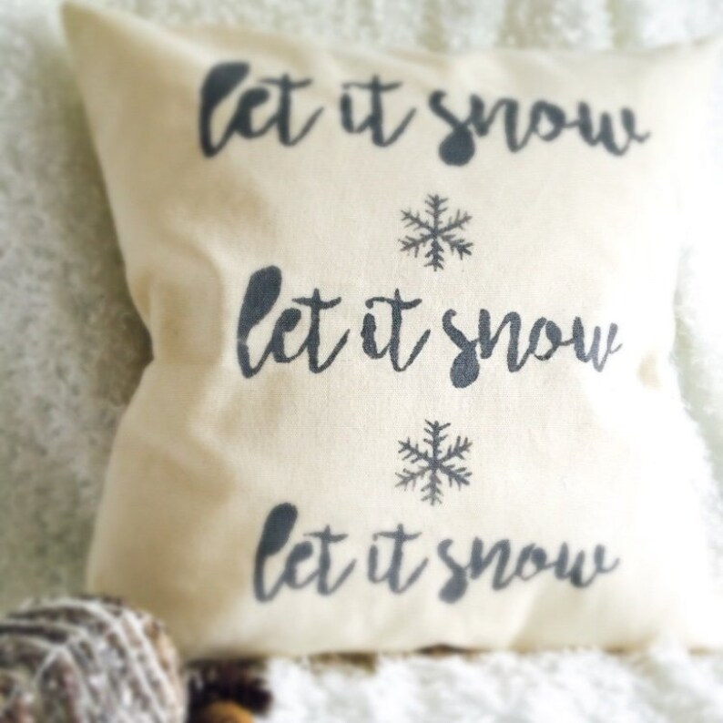 May include: A white throw pillow with the text "Let it snow" repeated three times in black lettering. Each line of text is accompanied by a black snowflake.