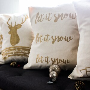 May include: Four decorative throw pillows with gold lettering and designs on a white background. The pillows feature the phrases "let it snow" and "merry christmas" with a reindeer and a Christmas tree design.