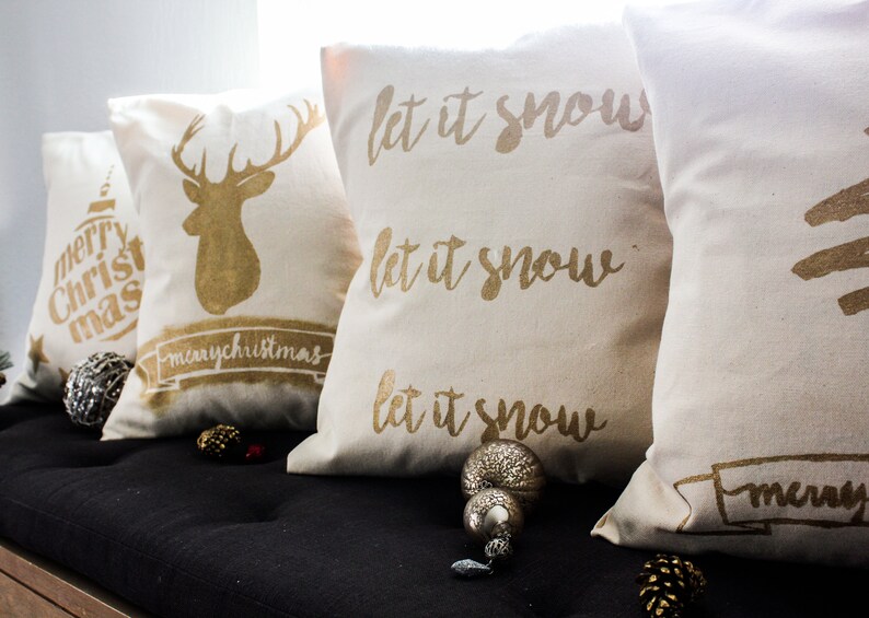 May include: Four decorative throw pillows with gold lettering and designs on a white background. The pillows feature the phrases "let it snow" and "merry christmas" with a reindeer and a Christmas tree design.