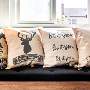 May include: Four decorative throw pillows with a Christmas theme. The pillows are beige with black lettering and designs. The pillows feature the text "merry christmas", a deer silhouette, and the text "let it snow" repeated three times. One pillow features a stylized Christmas tree.