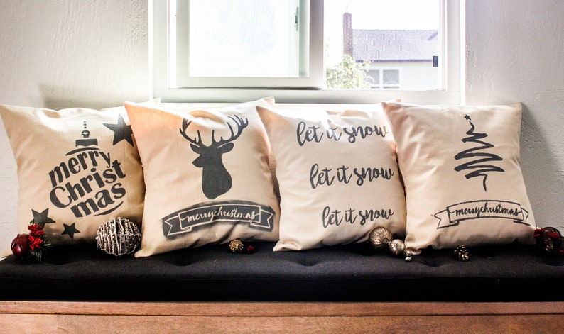 May include: Four decorative throw pillows with a Christmas theme. The pillows are beige with black lettering and designs. The pillows feature the text "merry christmas", a deer silhouette, and the text "let it snow" repeated three times. One pillow features a stylized Christmas tree.