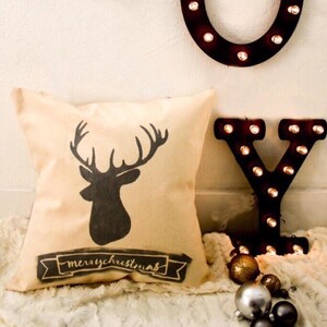May include: A beige throw pillow with a black silhouette of a reindeer and the text "merrychristmas" on a banner. The pillow is on a white faux fur surface with a black metal letter "Y" with light bulbs behind it.