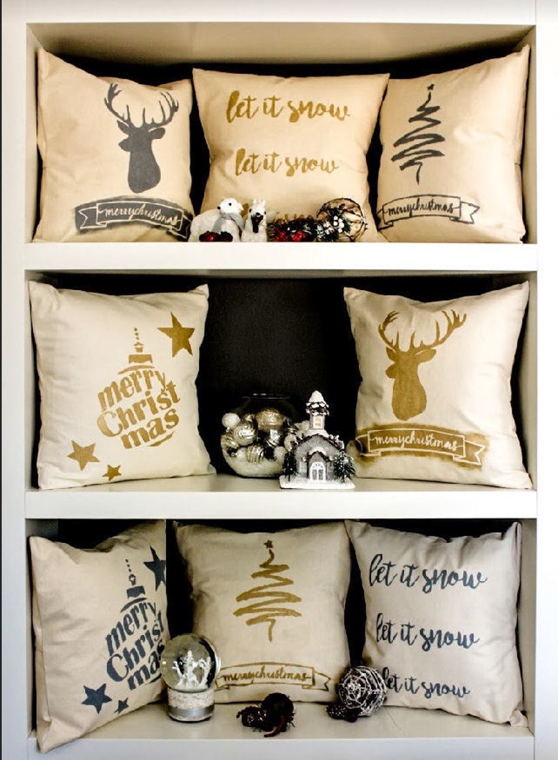 May include: A shelf with nine decorative throw pillows featuring Christmas themes. The pillows are beige with gold and black lettering and designs. The designs include a Christmas tree, a reindeer, and the words "let it snow" and "merry christmas".