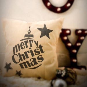 May include: A beige throw pillow with a black and white design of a Christmas ornament with the text "merry Christ mas" and two stars.