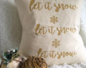 Puede incluir: Una almohada blanca con el texto "Let it snow" repetido tres veces en letras doradas con purpurina. Cada frase "Let it snow" va acompañada de un copo de nieve dorado.