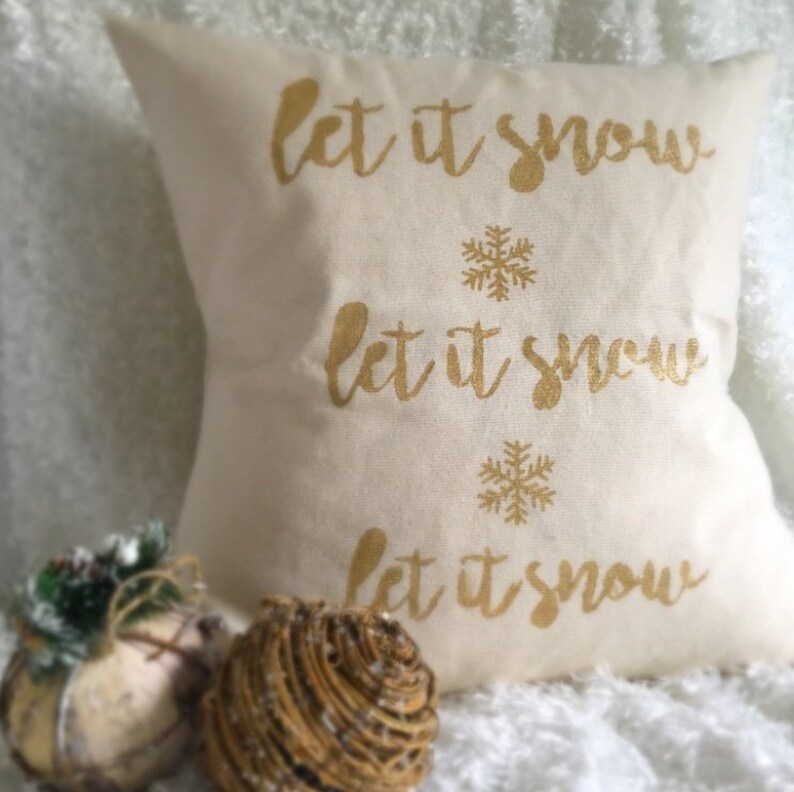 May include: A white throw pillow with the text "Let it snow" repeated three times in gold glitter lettering. Each "Let it snow" phrase is accompanied by a gold snowflake.