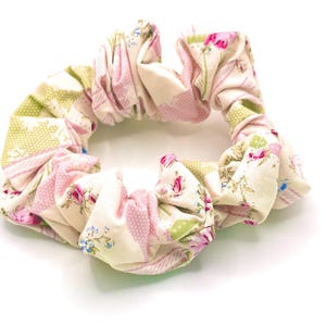 Puede incluir: Una scrunchie floral con un diseño de patchwork. Presenta rosas rosas, hojas verdes y un fondo crema. La tela está fruncida y doblada, creando una forma circular texturizada. Este accesorio para el cabello añade un toque vintage.