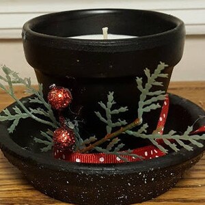 May include: Black terracotta pot with a white candle inside, decorated with a red and white ribbon and a green sprig with red berries. The pot is sitting on a black saucer.