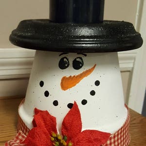 Puede incluir: Una maceta de flores pintada de blanco decorada para parecer un muñeco de nieve con un sombrero negro, una nariz de zanahoria naranja y ojos negros. El muñeco de nieve lleva una cinta a cuadros rojos y blancos con una flor de poinsettia roja.
