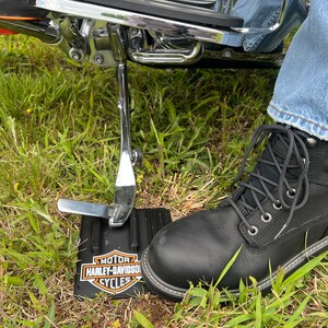 Puede incluir: Bota de trabajo de cuero negro con cordones grises y suela cosida blanca. La bota est&aacute; junto a un caballete de motocicleta negro y un logotipo de Harley-Davidson sobre una base negra, apoyada sobre hierba.