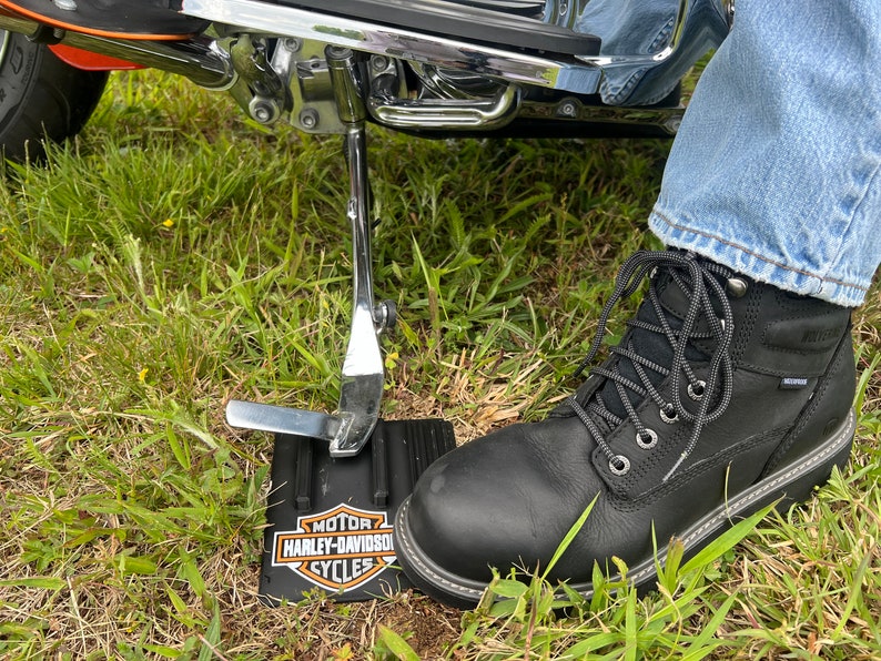 Puede incluir: Bota de trabajo de cuero negro con cordones grises y suela cosida blanca. La bota est&aacute; junto a un caballete de motocicleta negro y un logotipo de Harley-Davidson sobre una base negra, apoyada sobre hierba.
