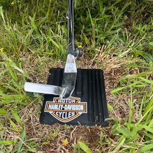 Puede incluir: Un caballete negro para motocicleta con acabado cromado, apoyado sobre una base negra con el logotipo de Harley-Davidson Cycles en naranja y blanco. El caballete est&aacute; en la hierba.