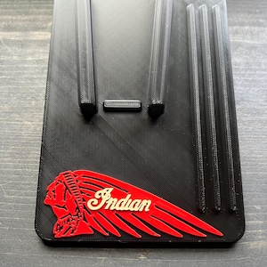 Puede incluir: Soporte para tel&eacute;fono negro impreso en 3D con el logotipo de Indian Motorcycle en rojo y crema. El soporte tiene dos soportes verticales y tres ranuras verticales. El soporte rectangular se encuentra sobre una superficie de madera.