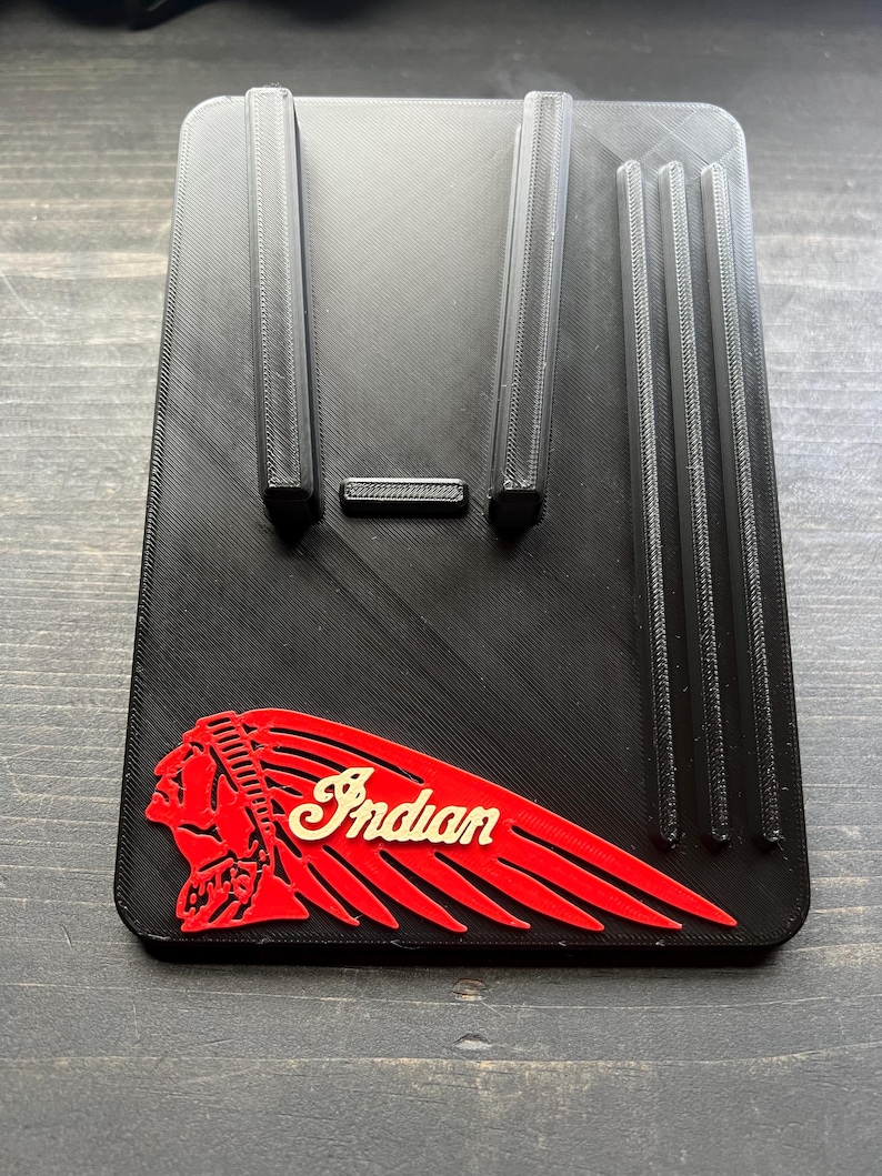 Puede incluir: Soporte para tel&eacute;fono negro impreso en 3D con el logotipo de Indian Motorcycle en rojo y crema. El soporte tiene dos soportes verticales y tres ranuras verticales. El soporte rectangular se encuentra sobre una superficie de madera.