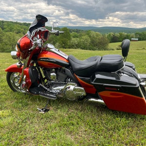 Puede incluir: Una motocicleta Harley-Davidson roja y naranja estacionada en un campo cubierto de hierba. La motocicleta tiene detalles cromados, un asiento negro y alforjas. El fondo presenta &aacute;rboles verdes y un cielo nublado.