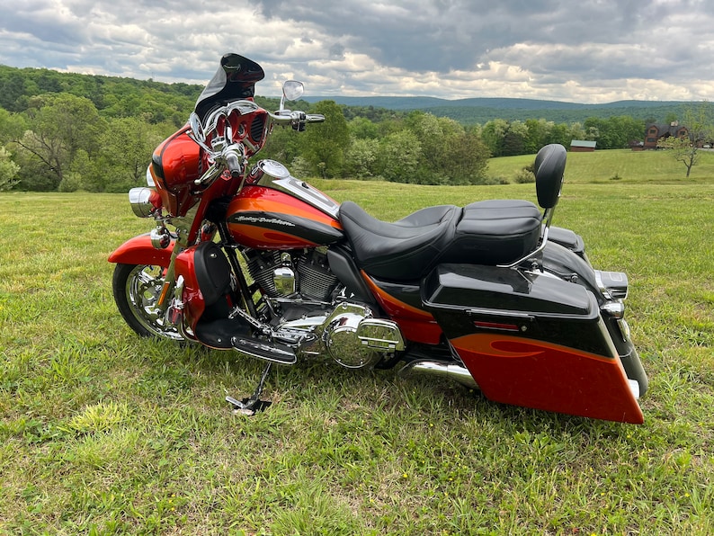 Puede incluir: Una motocicleta Harley-Davidson roja y naranja estacionada en un campo cubierto de hierba. La motocicleta tiene detalles cromados, un asiento negro y alforjas. El fondo presenta &aacute;rboles verdes y un cielo nublado.