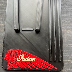 Puede incluir: Accesorio rectangular negro para motocicleta con la palabra "Indian" en escritura color crema y un logotipo de cabeza de nativo americano rojo. El accesorio tiene crestas verticales y horizontales y una superficie texturizada.