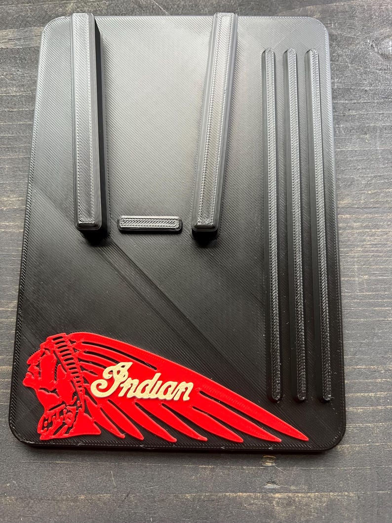 Puede incluir: Accesorio rectangular negro para motocicleta con la palabra "Indian" en escritura color crema y un logotipo de cabeza de nativo americano rojo. El accesorio tiene crestas verticales y horizontales y una superficie texturizada.