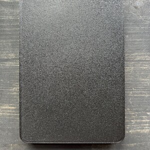 Puede incluir: Un objeto rectangular negro texturizado con esquinas redondeadas. La superficie tiene un aspecto moteado. El art&iacute;culo est&aacute; colocado sobre un fondo de vetas de madera oscura. El objeto parece ser una tapa o una bandeja peque&ntilde;a.