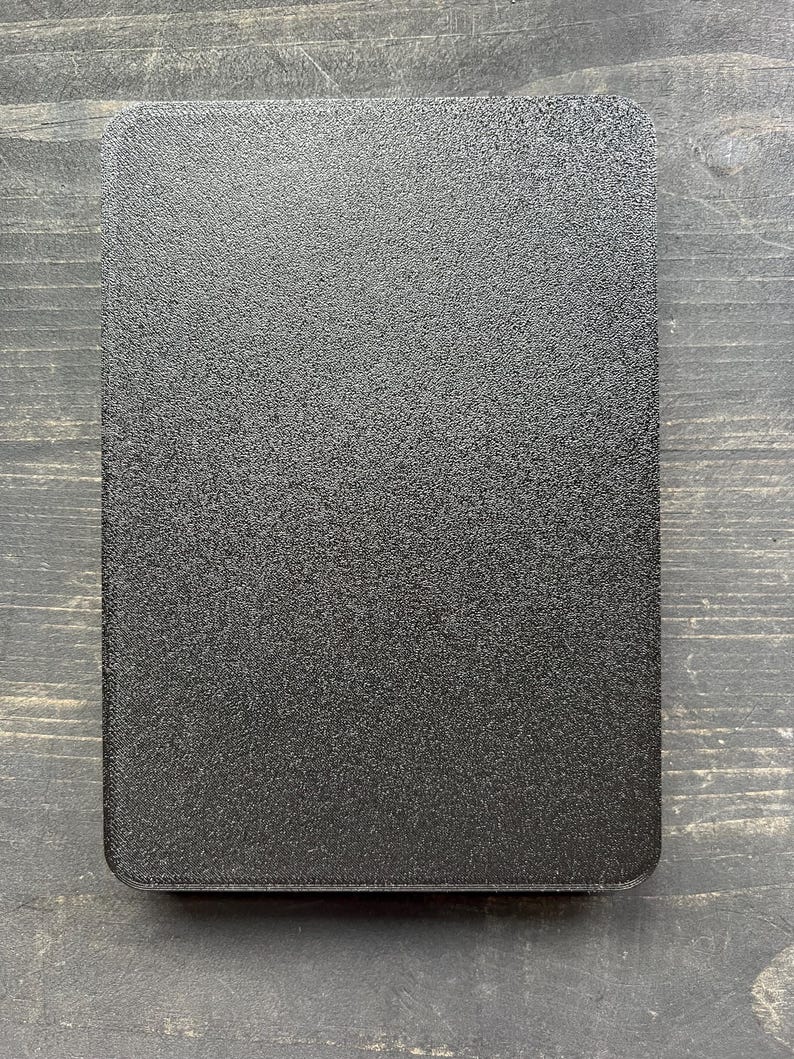 Puede incluir: Un objeto rectangular negro texturizado con esquinas redondeadas. La superficie tiene un aspecto moteado. El art&iacute;culo est&aacute; colocado sobre un fondo de vetas de madera oscura. El objeto parece ser una tapa o una bandeja peque&ntilde;a.