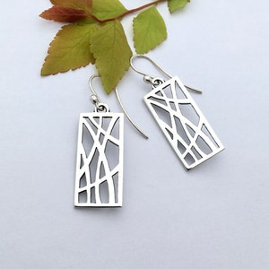 May include: Two silver rectangular earrings with an open, abstract design. The earrings feature intersecting lines, creating a geometric pattern. The earrings have a hook closure and are displayed on a white surface with a green and red leaf branch.