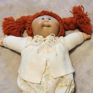 May include: A vintage Cabbage Patch Kid doll with red yarn pigtails, blue eyes, and a soft, fabric body. The doll wears a white knit sweater, floral print bloomers with ruffled trim, and has a sweet, smiling expression.