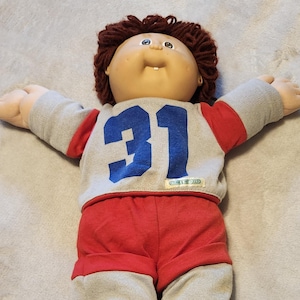 May include: A vintage Cabbage Patch Kids doll, wearing a gray and red outfit with the number 31 in blue on the shirt. The doll has brown yarn hair, a round face, and open arms.