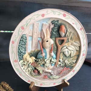 May include: A ceramic plate with a 3D design of Peter Rabbit holding carrots in a garden setting. The plate is decorated with a pink and green border and features a robin perched on a rake.