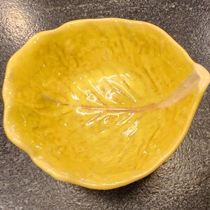 May include: A yellow ceramic leaf-shaped dish. The dish has a glossy finish and a detailed vein pattern. The edges are slightly raised, and the overall shape resembles a leaf. This decorative dish is suitable for serving small snacks or as a decorative accent.