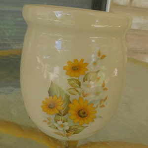 May include: A cream-colored ceramic vase with a floral design. The vase features yellow and white flowers with green leaves.