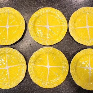 May include: Six yellow ceramic plates with a lemon slice design. Each plate features a bright yellow color with white accents, and a textured surface. The plates are arranged in two rows of three.