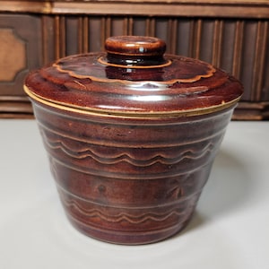 May include: A brown ceramic jar with a lid. The jar has a round shape with wavy and linear patterns. The lid has a scalloped edge and a small knob. The jar is a dark brown color.