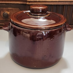May include: A small, round, brown ceramic pot with a matching lid. The pot has two small handles on either side and a glossy finish. The lid has a small knob on top for easy removal. The pot is likely used for cooking or storage.